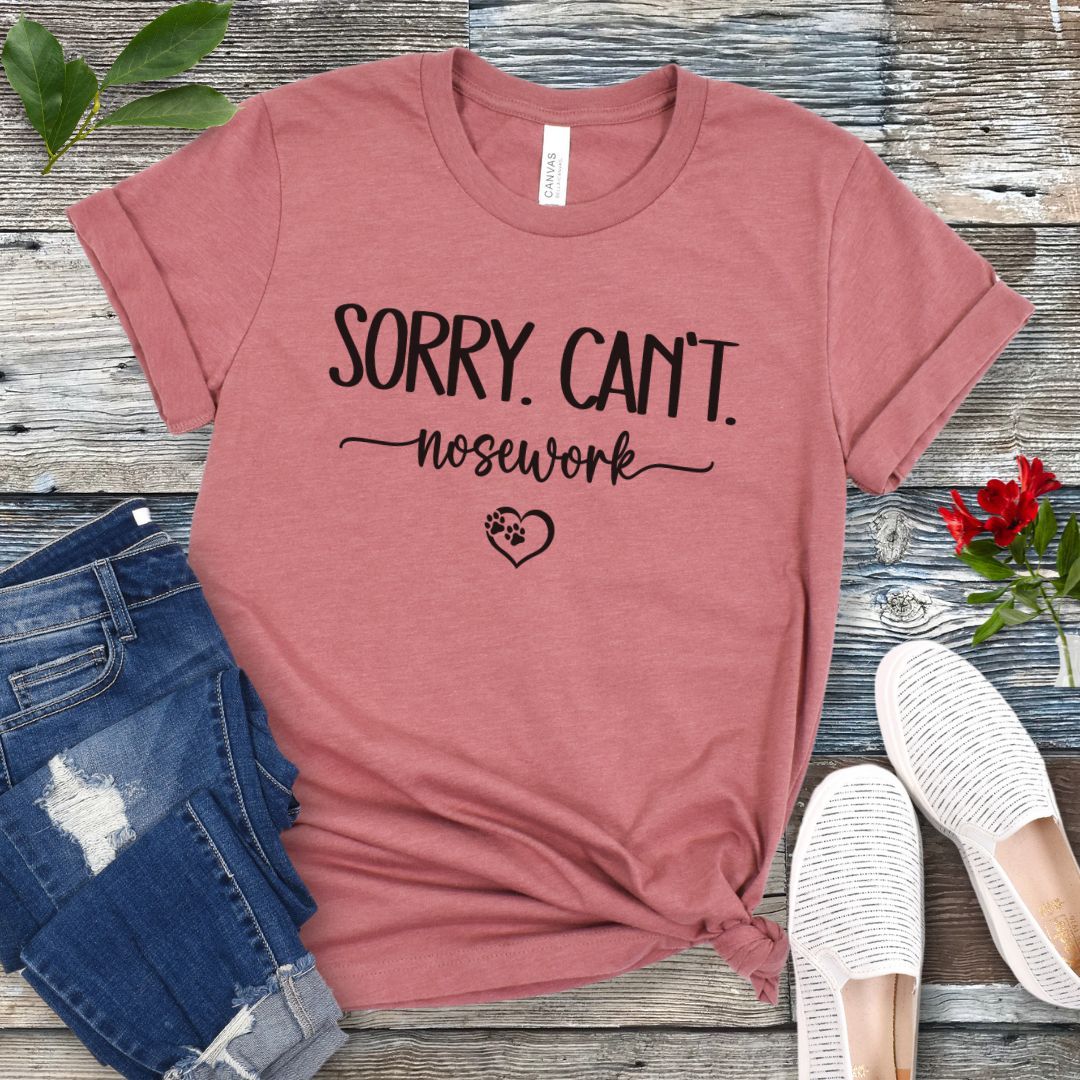 Mauve dog sports t-shirt with 'Sorry. Can't. Nosework' text on a wooden surface with jeans and shoes.