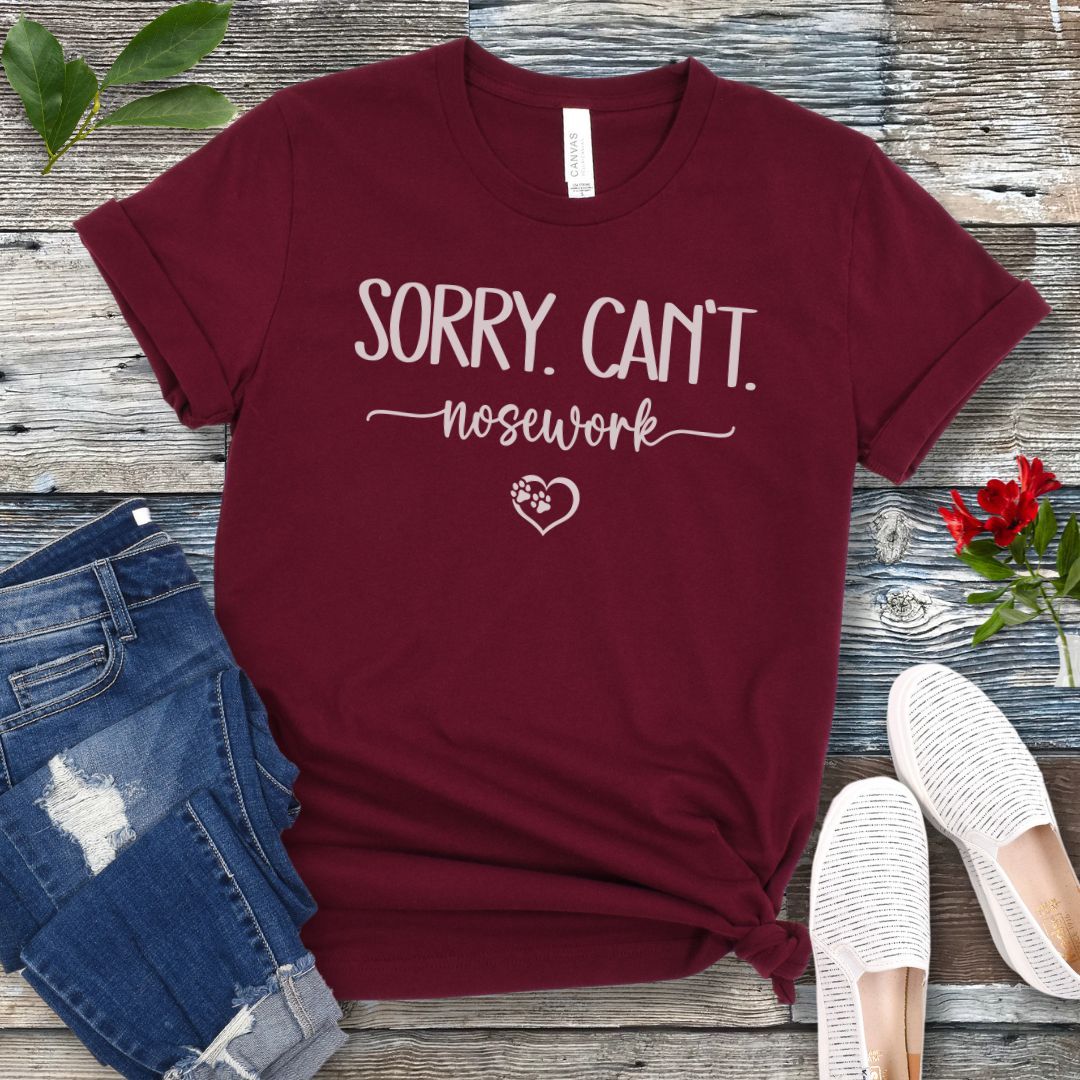 Maroon dog sports t-shirt with 'Sorry. Can't. Nosework' text on a wooden surface with jeans and shoes.