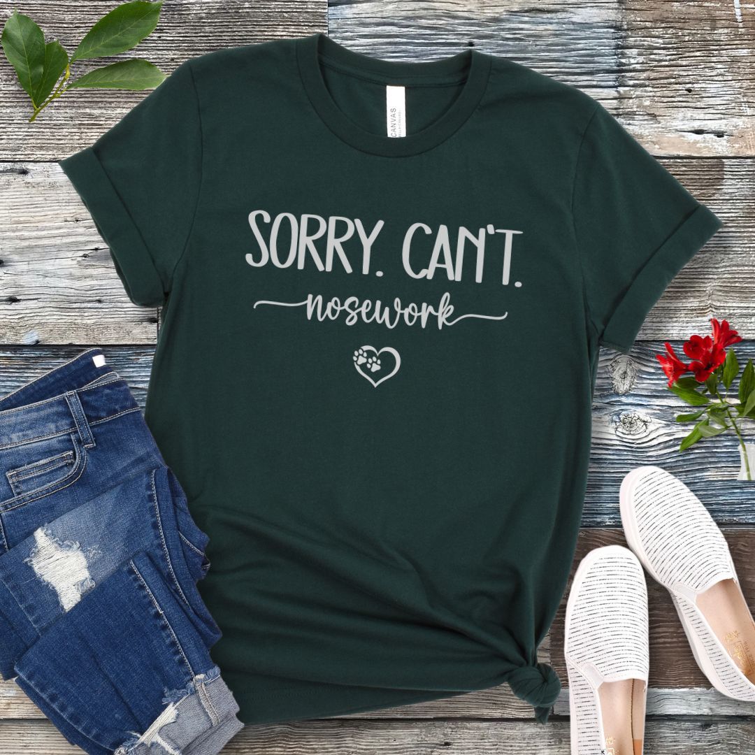 Dark green dog sports t-shirt with 'Sorry. Can't. Nosework' text on a wooden surface with jeans and shoes.