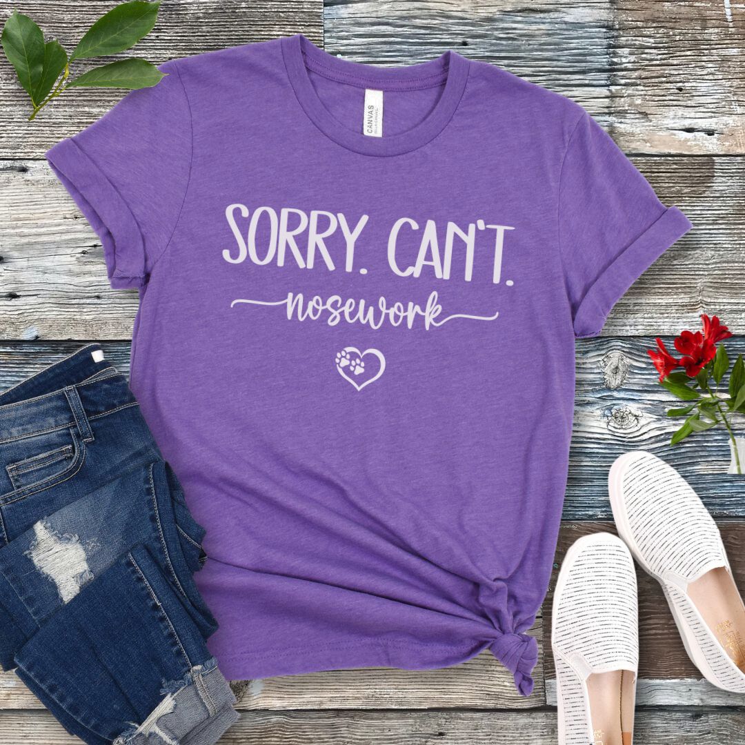 Purple dog sports t-shirt with 'Sorry. Can't. Nosework' text on a wooden surface with jeans and shoes.