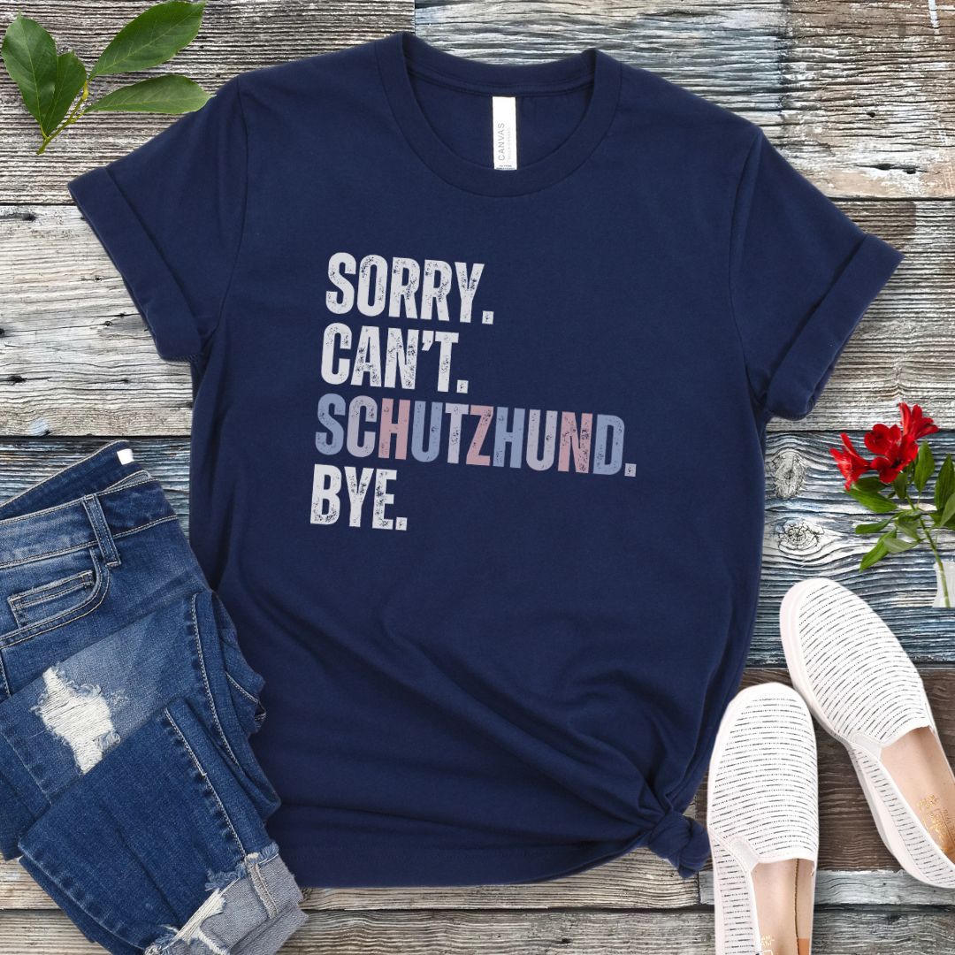 Navy blue t-shirt with 'SORRY. CAN'T. SCHUTZHUND. BYE' in colorful text, presented on a wooden surface with jeans and shoes.