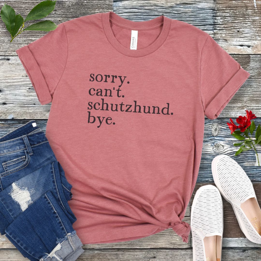 Mauve t-shirt with text 'sorry. can't. schutzhund. bye.' on a wooden surface with jeans and shoes.