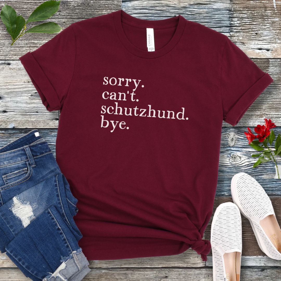 Maroon t-shirt with text 'sorry. can't. schutzhund. bye.' on a wooden surface with jeans and shoes.
