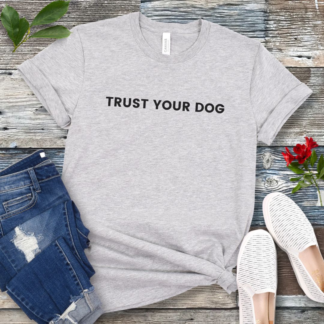 Light grey dog training quote t-shirt with text 'Trust Your Dog' on a wooden surface with jeans and shoes.