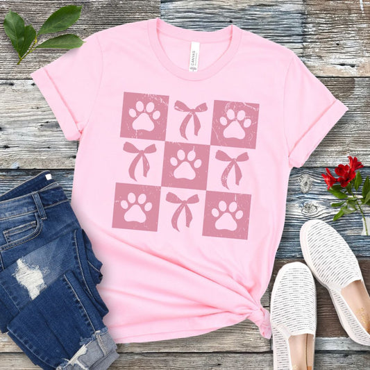 A pink dog lover t-shirt with a checkered pattern featuring rose-colored paw prints and bows, paired with blue jeans and white sneakers.