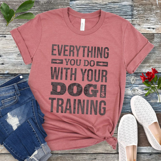 Mauve t-shirt with the text 'EVERYTHING YOU DO WITH YOUR DOG IS TRAINING' text design, displayed flat with a pair of blue jeans and white shoes.