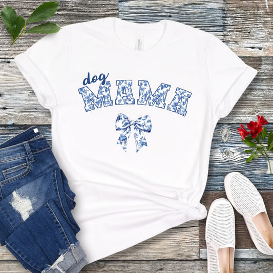 A white t-shirt with 'Dog Mama' written in a vintage-inspired floral print, displayed flat with a pair of blue jeans, white shoes, and accessories.
