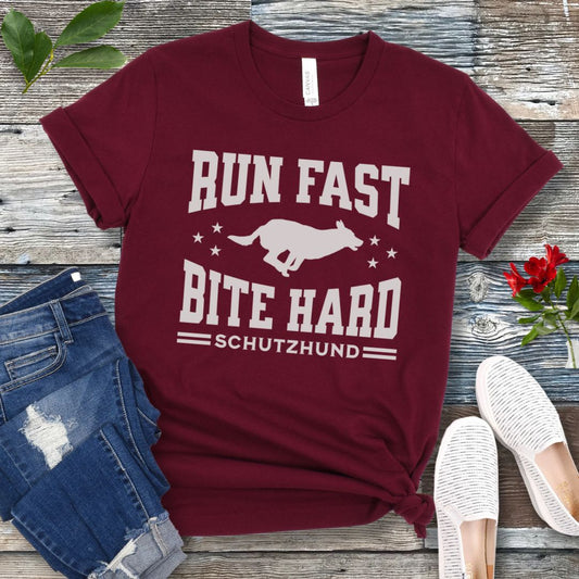 A maroon unisex IGP/Schutzhund T-shirt with running dog and the text 'RUN FAST BITE HARD SCHUTZHUND' printed in white, displayed flat on a wooden surface with denim jeans and white shoes beside it.