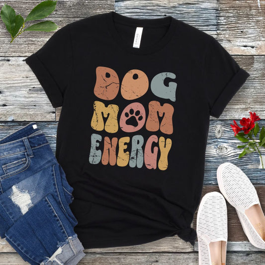 A black t-shirt for dog lovers with 'DOG MOM ENERGY' printed in multicolor retro text, placed on a wooden surface with a pair of blue jeans and white shoes in the background.