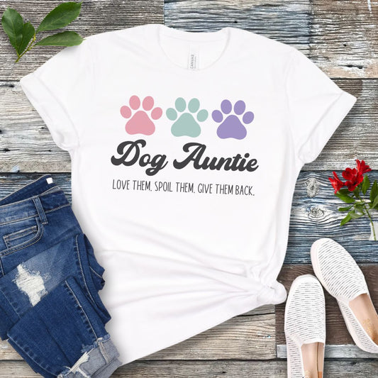 A white dog lover t-shirt with 'Dog Auntie, Love Them Spoil Them, Give Theme Back' text and colorful paw prints design, placed on a wooden surface with jeans and white shoes in the background.