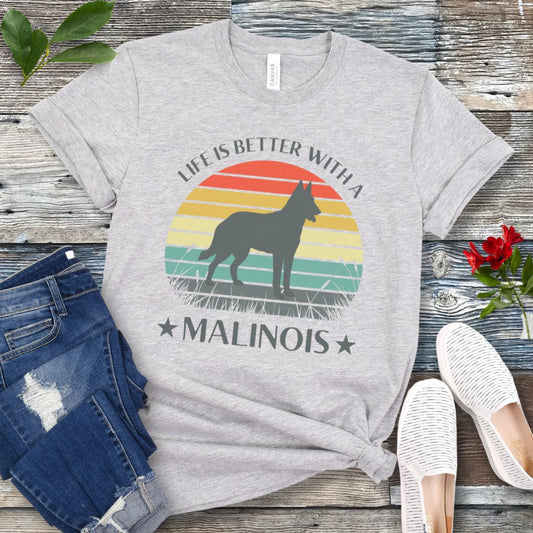 A grey Belgian Malinois t-shirt with a retro sunset graphic, a Malinois silhouette, and the text 'LIFE IS BETTER WITH A MALINOIS', displayed on a wooden surface with denim jeans and white shoes in the background.