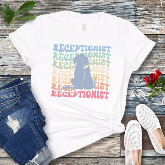 A white veterinary receptionist t-shirt with the text 'RECEPTIONIST' in a wavy, retro, repeating gradient design, and a puppy and kitten silhouette over the top. Displayed on a wooden surface with a pair of blue jeans and white shoes in the background.