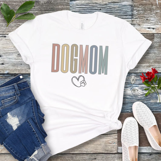 White dog mom t-shirt with modern multicolor “DOG MOM” text and heart paw graphic, styled flat lay with jeans and slip-on shoes; minimalist dog lover shirt and gift for dog moms.