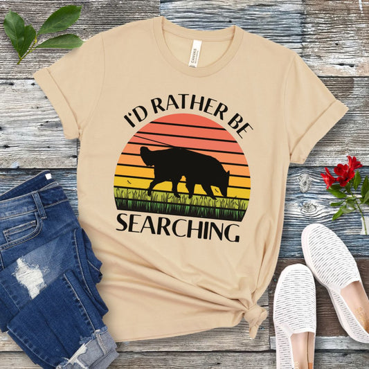 Cream nosework t-shirt with custom breed sniffing dog graphic and 'I'd Rather Be Searching' text on a wooden surface with jeans and shoes.