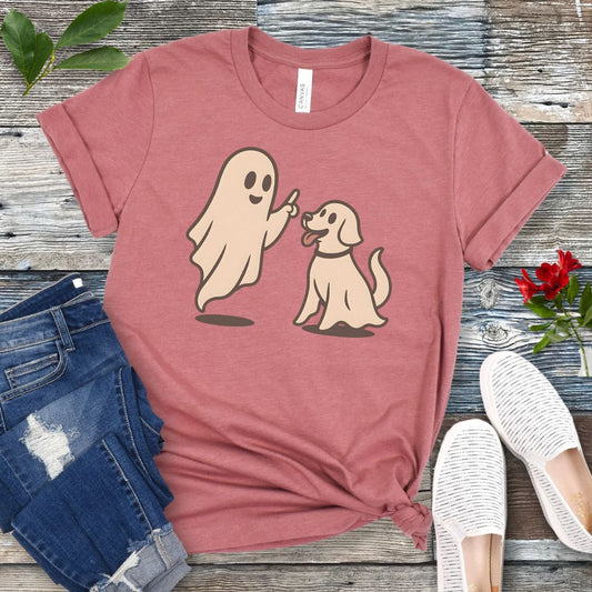 A mauve dog trainer t-shirt with a graphic of a ghost training a dog, placed on a wooden surface with denim jeans and white shoes in the background.