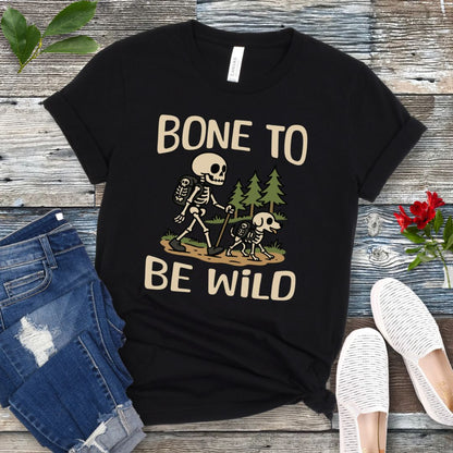 A black t-shirt with a graphic print of a skeleton and a dog on a trail, with text reading 'BONE TO BE WILD'. The t-shirt is presented flat with a pair of blue jeans and white shoes in the background.