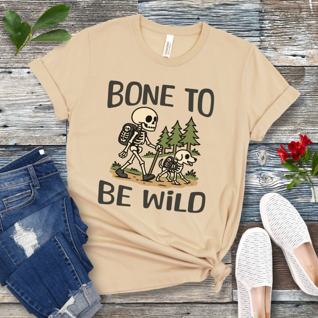A beige-colored t-shirt with a graphic print of a skeleton and a dog on a trail, with text reading 'BONE TO BE WILD'. The t-shirt is presented flat with a pair of blue jeans and white shoes in the background.