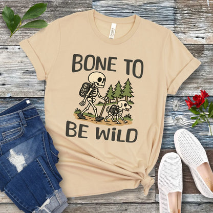 A beige-colored t-shirt with a graphic print of a skeleton and a dog on a trail, with text reading 'BONE TO BE WILD'. The t-shirt is presented flat with a pair of blue jeans and white shoes in the background.