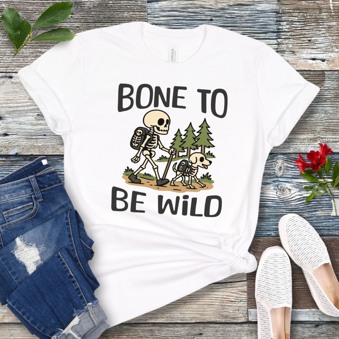 A white t-shirt with a graphic print of a skeleton and a dog on a trail, with text reading 'BONE TO BE WILD'. The t-shirt is presented flat with a pair of blue jeans and white shoes in the background.