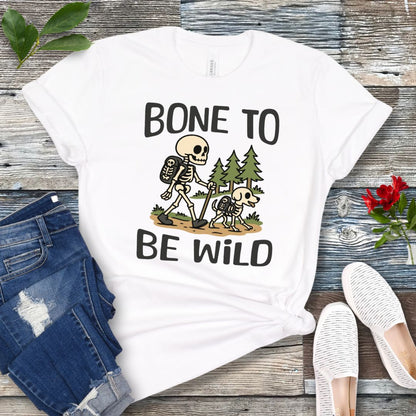 A white t-shirt with a graphic print of a skeleton and a dog on a trail, with text reading 'BONE TO BE WILD'. The t-shirt is presented flat with a pair of blue jeans and white shoes in the background.