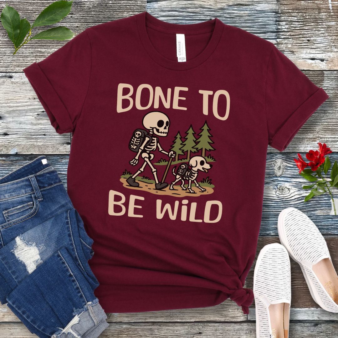 A maroon t-shirt with a graphic print of a skeleton and a dog on a trail, with text reading 'BONE TO BE WILD'. The t-shirt is presented flat with a pair of blue jeans and white shoes in the background.