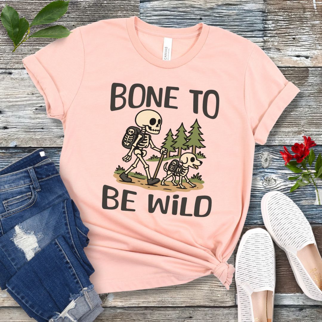 A peach-colored t-shirt with a graphic print of a skeleton and a dog on a trail, with text reading 'BONE TO BE WILD'. The t-shirt is presented flat with a pair of blue jeans and white shoes in the background.
