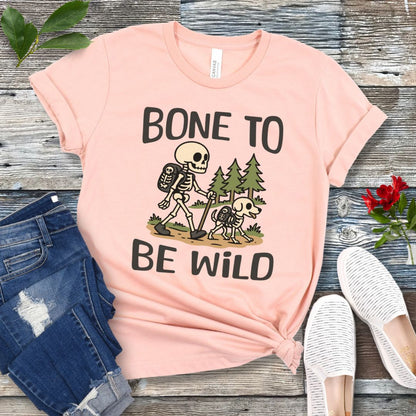 A peach-colored t-shirt with a graphic print of a skeleton and a dog on a trail, with text reading 'BONE TO BE WILD'. The t-shirt is presented flat with a pair of blue jeans and white shoes in the background.