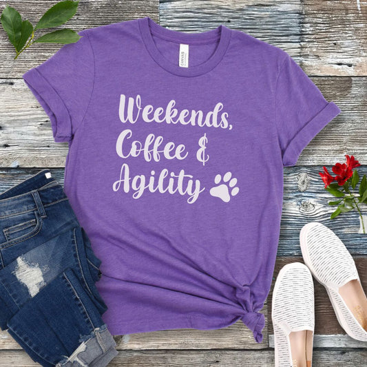 Purple dog sports t-shirt with 'Weekends, Coffee & Agility' text on a wooden surface with jeans and shoes.