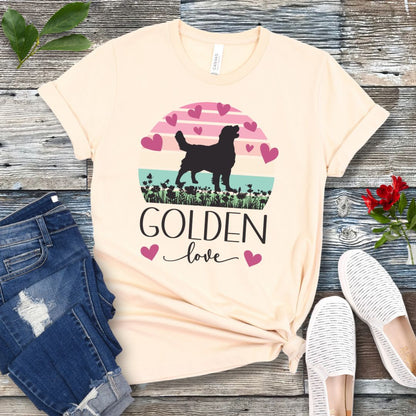 Beige t-shirt with a graphic of a Golden Retriever surrounded by hearts and a vintage sunset, and 'Golden Love' text below, presented on a wooden surface with jeans and shoes.