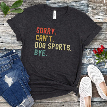 Dark grey t-shirt with the text 'SORRY, CAN'T, DOG SPORTS, BYE' printed in colorful letters on a wooden background, accompanied by casual clothing and shoes.
