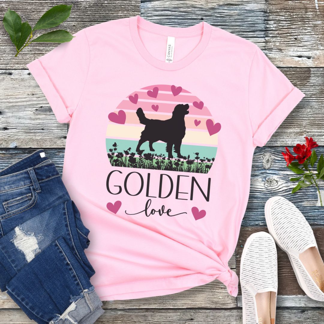 Pink dog mom t-shirt with a graphic of a Golden Retriever surrounded by hearts and a vintage sunset, and 'Golden Love' text below, presented on a wooden surface with jeans and shoes.