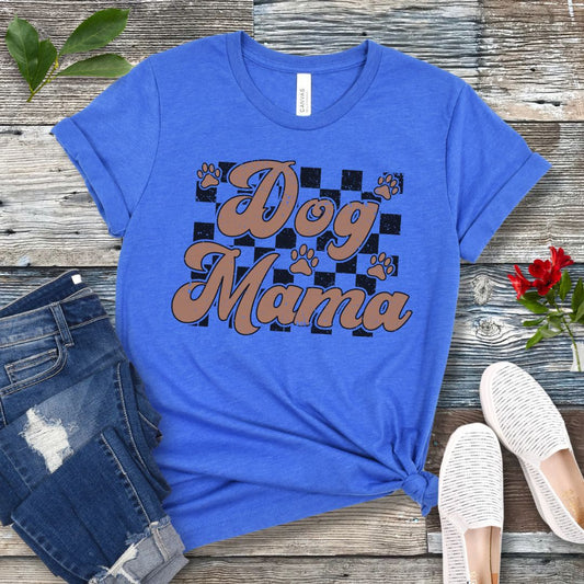 A royal blue unisex short-sleeve t-shirt with 'Dog Mama' text in retro style lettering and paw prints over a black checkerboard pattern, laid out flat with a pair of blue jeans and white shoes on a wooden surface.
