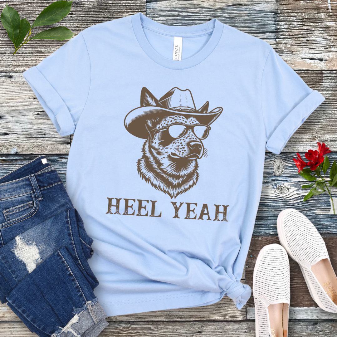 Light blue t-shirt with a graphic of a Cattle dog wearing sunglasses and a cowboy hat, text 'HEEL YEAH', displayed on a wooden surface with jeans and shoes.