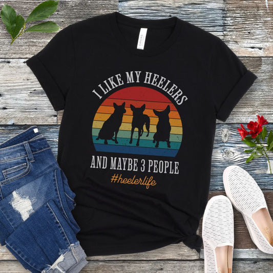 Black t-shirt with vintage sunset graphic, 3 Heeler/Cattle Dog silhouettes, and the text 'I Like My Heelers and maybe 3 people. #heelerlife' on a wooden surface with jeans and shoes.