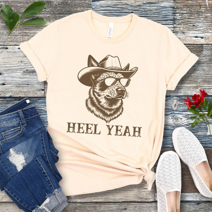Beige t-shirt with a graphic of a Cattle dog wearing sunglasses and a cowboy hat, text 'HEEL YEAH', displayed on a wooden surface with jeans and shoes.