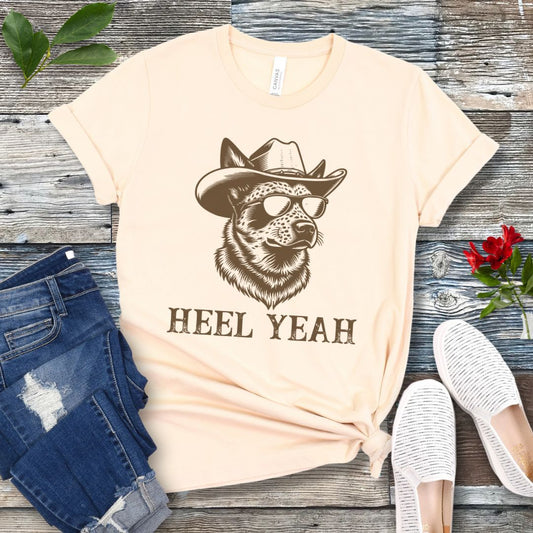 Beige t-shirt with a graphic of a Cattle dog wearing sunglasses and a cowboy hat, text 'HEEL YEAH', displayed on a wooden surface with jeans and shoes.