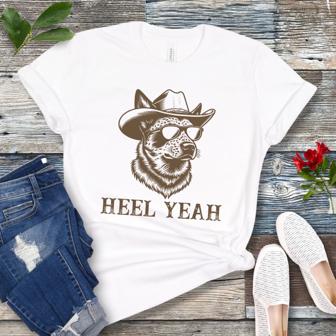 White t-shirt with a graphic of a Cattle dog wearing sunglasses and a cowboy hat, text 'HEEL YEAH', displayed on a wooden surface with jeans and shoes.