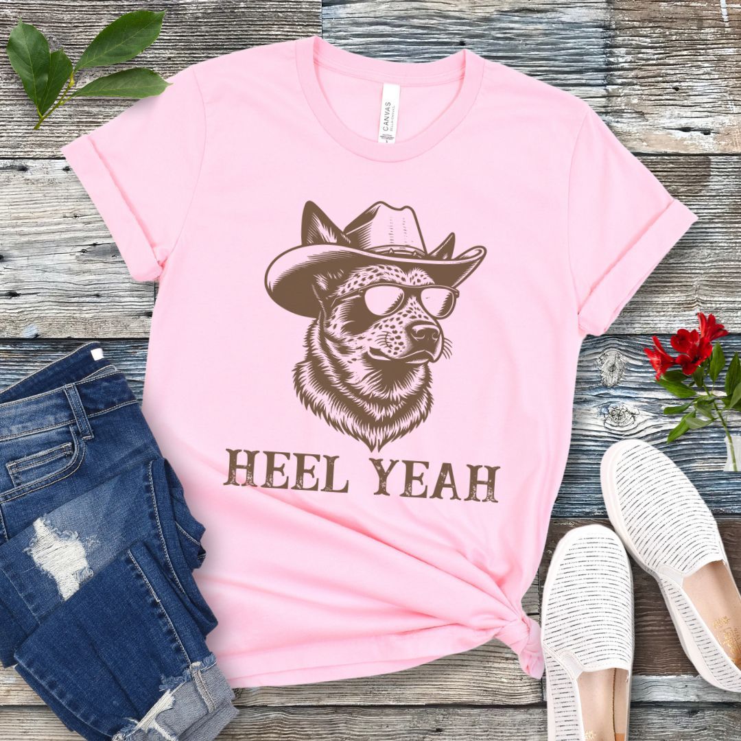 Pink t-shirt with a graphic of a Cattle dog wearing sunglasses and a cowboy hat, text 'HEEL YEAH', displayed on a wooden surface with jeans and shoes.
