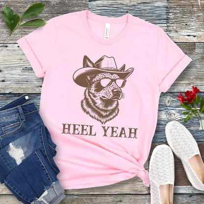 Pink t-shirt with a graphic of a Cattle dog wearing sunglasses and a cowboy hat, text 'HEEL YEAH', displayed on a wooden surface with jeans and shoes.