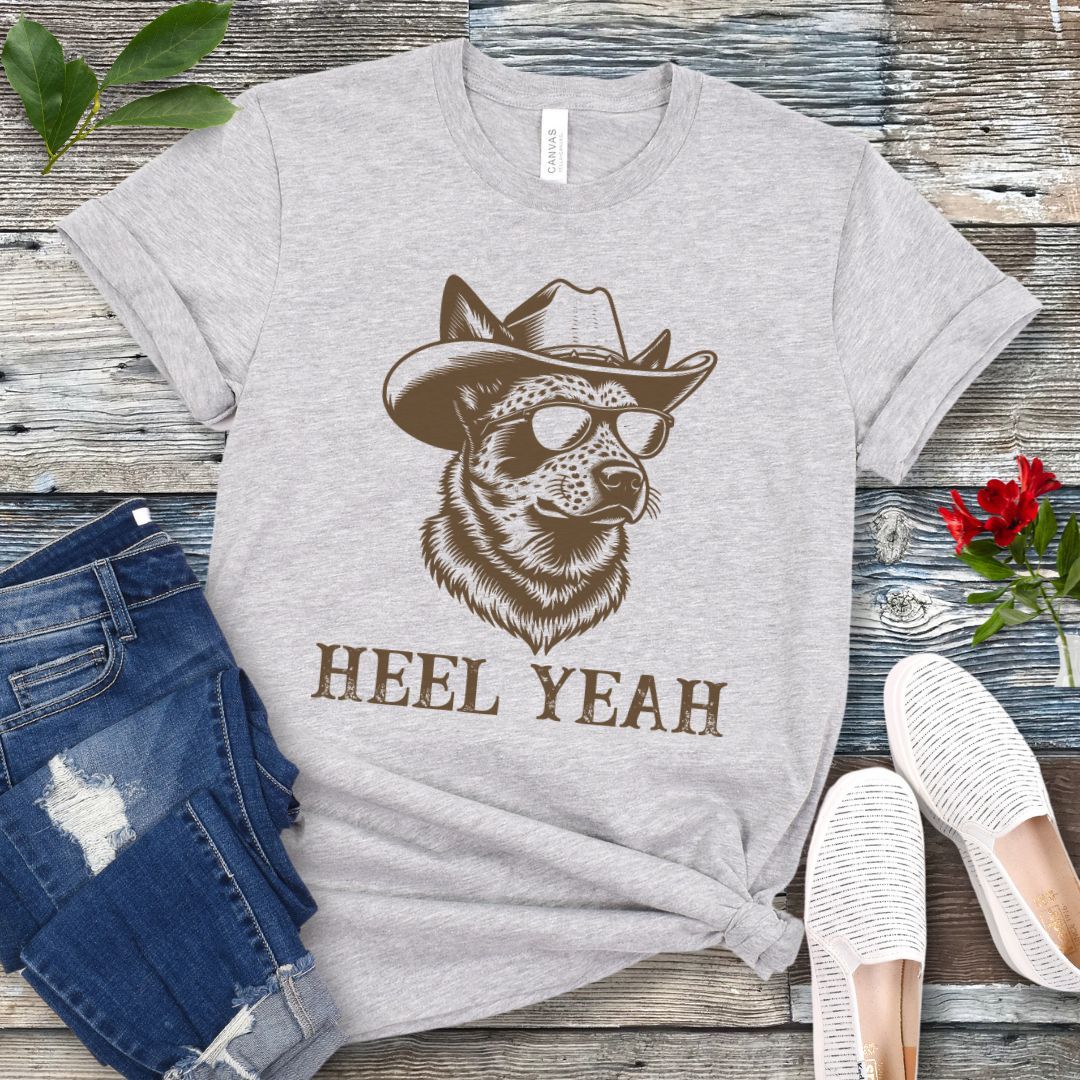 Light grey t-shirt with a graphic of a Cattle dog wearing sunglasses and a cowboy hat, text 'HEEL YEAH', displayed on a wooden surface with jeans and shoes.