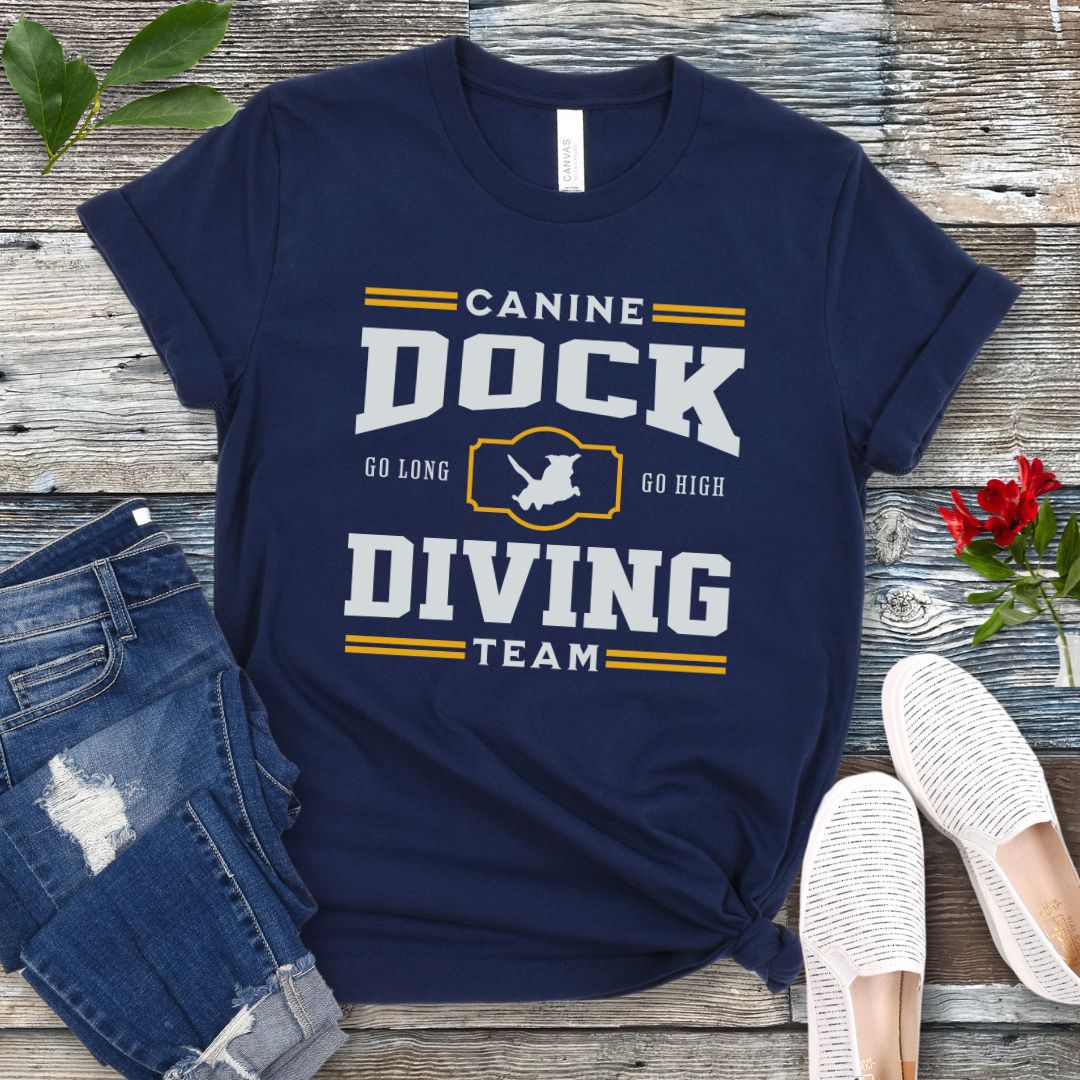 A navy blue t-shirt with 'CANINE DOCK DIVING TEAM' text and a graphic of a dock jumping dog, placed on a wooden surface with jeans and white shoes in the background.