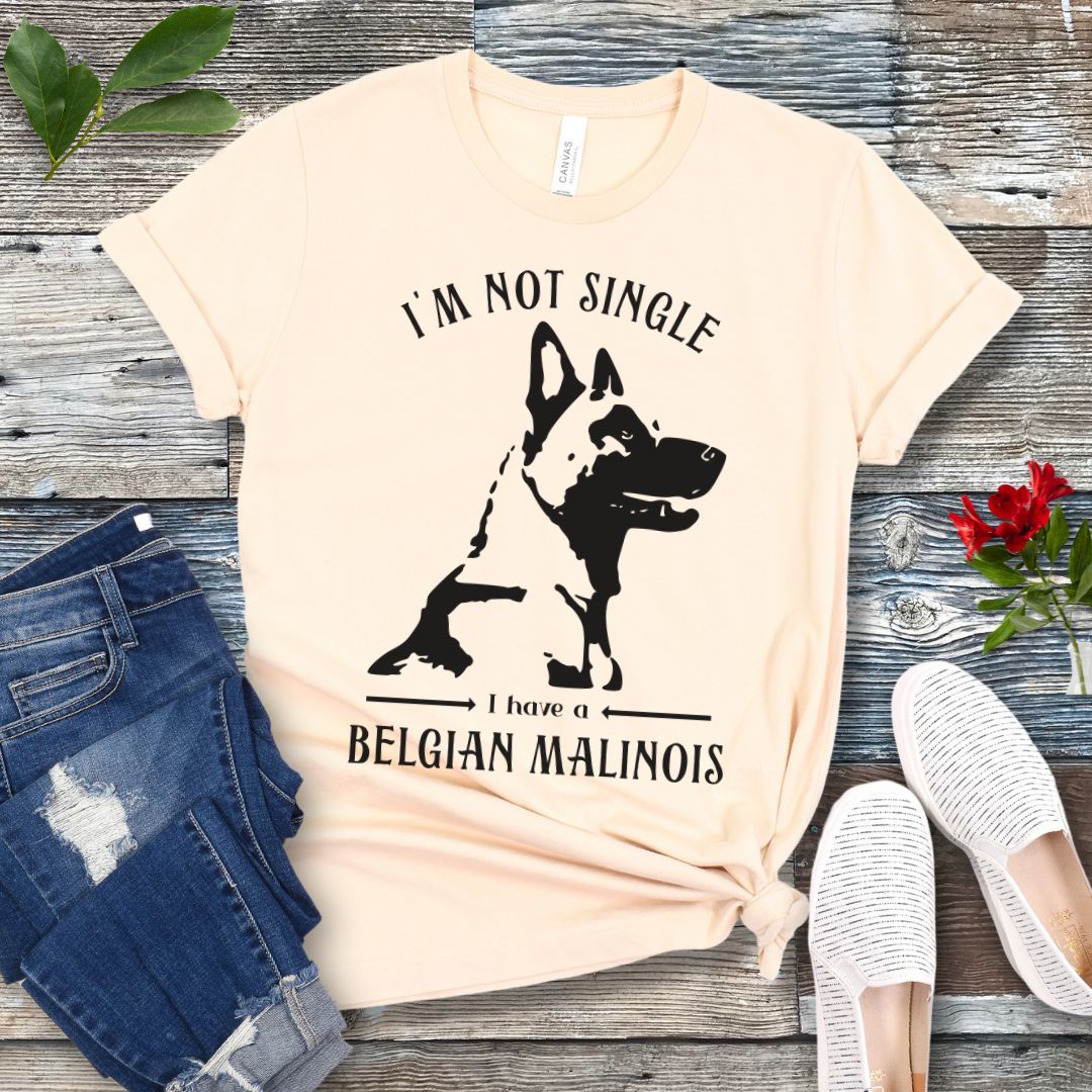 Beige t-shirt with "I'm Not Single I have a Belgian Malinois" text and silhouette of a Belgian Malinois dog printed on it, displayed flat with a pair of jeans and white shoes on a wooden surface.