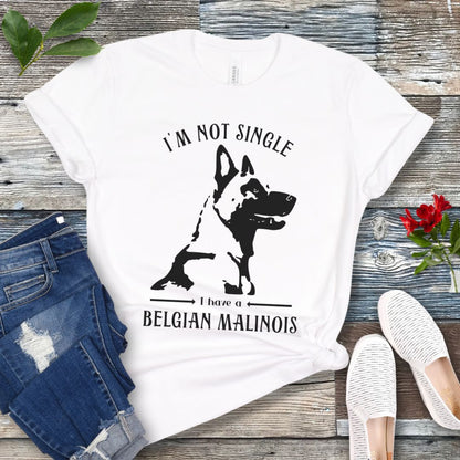 White t-shirt with "I'm Not Single I have a Belgian Malinois" text and silhouette of a Belgian Malinois dog printed on it, displayed flat with a pair of jeans and white shoes on a wooden surface.
