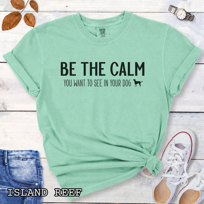 Light teal Comfort Colors t-shirt with text 'BE THE CALM YOU WANT TO SEE IN YOUR DOG' on a wooden surface with jeans and a plant.