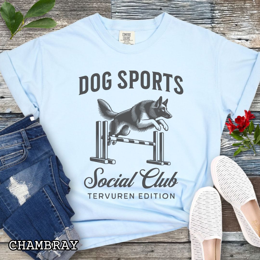Blue Comfort Colors t-shirt with illustration of a Belgian Tervuren jumping a bar jump, and the text 'Dog Sports Social Club, Tervuren Edition' in vintage style lettering, shown on a wooden surface.