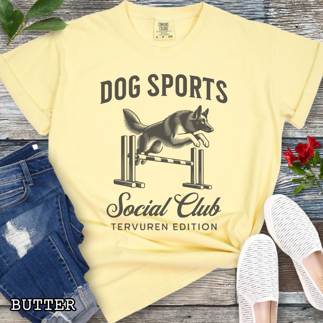 Yellow Comfort Colors t-shirt with illustration of a Belgian Tervuren jumping a bar jump, and the text 'Dog Sports Social Club, Tervuren Edition' in vintage style lettering, shown on a wooden surface.