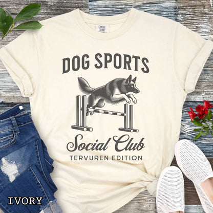 Beige Comfort Colors t-shirt with illustration of a Belgian Tervuren jumping a bar jump, and the text 'Dog Sports Social Club, Tervuren Edition' in vintage style lettering, shown on a wooden surface.