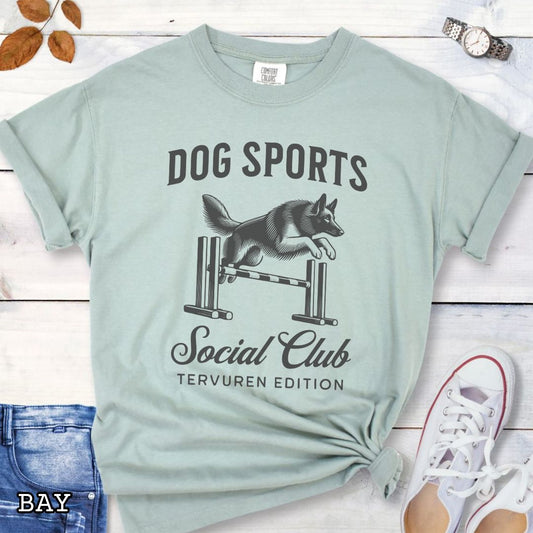 Bay green Comfort Colors t-shirt with illustration of a Belgian Tervuren jumping a bar jump, and the text 'Dog Sports Social Club, Tervuren Edition' in vintage style lettering, shown on a wooden surface.