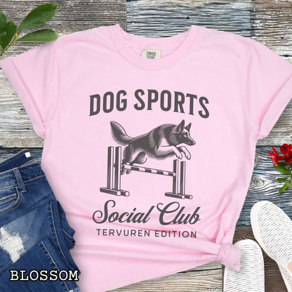 Pink Comfort Colors t-shirt with illustration of a Belgian Tervuren jumping a bar jump, and the text 'Dog Sports Social Club, Tervuren Edition' in vintage style lettering, shown on a wooden surface.