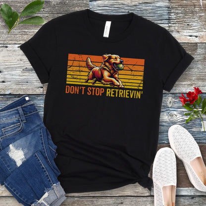 Black t-shirt with vintage sunset style horizontal stripes, a Golden Retriever running with a tennis ball, and the humorous text 'Don't Stop Retrieving', shown on a wooden surface with jeans and shoes.